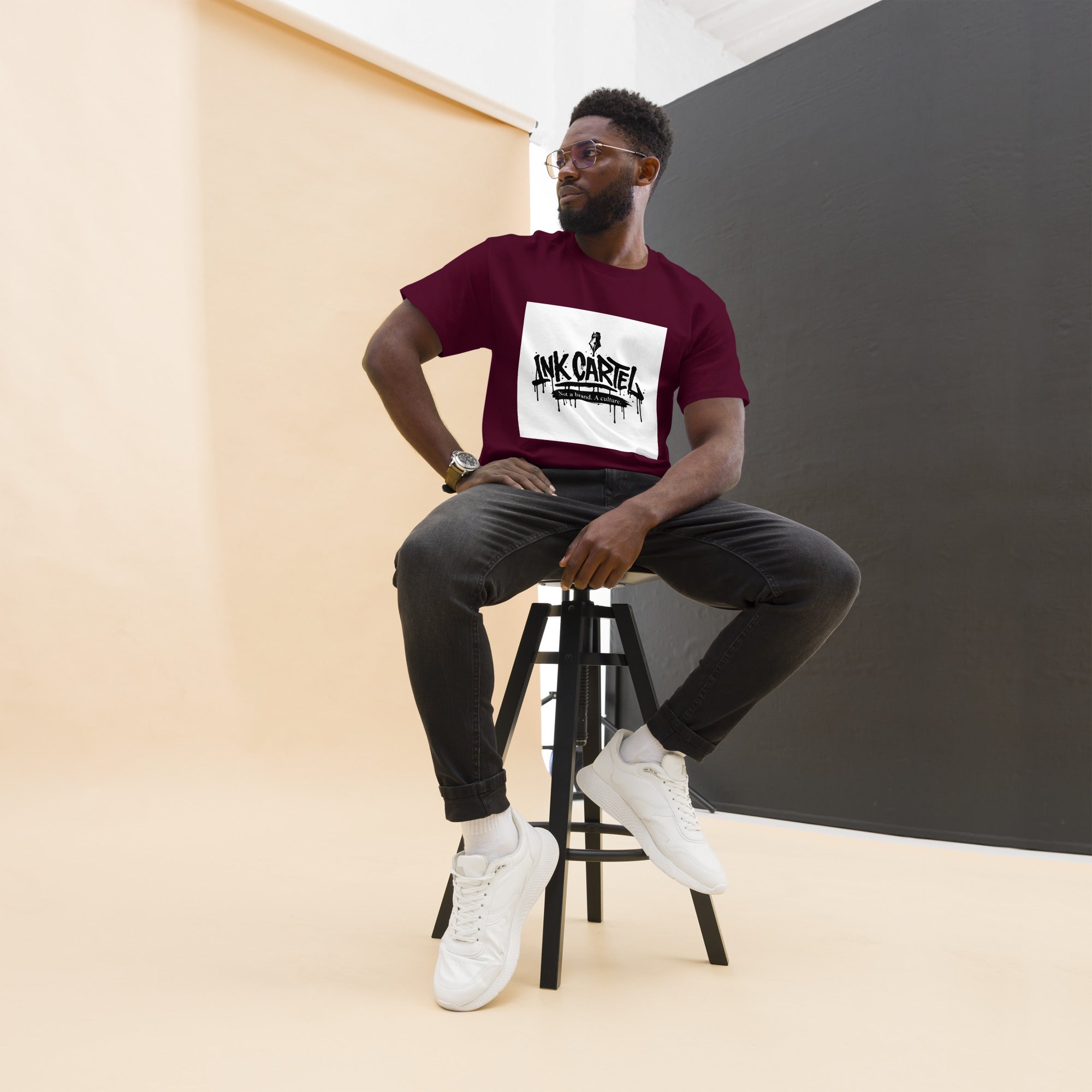 Man sitting on a stool wearing a maroon t-shirt with a logo, black pants, and white sneakers.